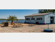 Ein einfaches Gebäude mit weißer Fassade und zahlreichen Fenstern steht am Ufer eines Sees. Davor befindet sich eine Fläche mit Sitzbänken, Tischen, einer großen Palme in einem Pflanzkübel und sandigem Boden, umgeben von einer betonierten Umgebung.