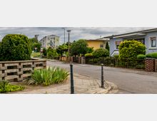 Straßenszene in einer Wohngegend mit Einfamilienhäusern, gepflegten Vorgärten und niedrigen Zäunen. Im Hintergrund ein Gebäude mit großem Wandgemälde, Straßenlaternen und Verkehrszeichen sind ebenfalls sichtbar.