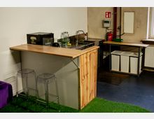 Kleine Küchenzeile mit hölzerner Theke und Spüle, daneben ein kleiner Kühlschrank und Abstellfläche mit Gläsern und Behältern. Im Hintergrund befinden sich Mülleimer, ein Feuerlöscher und ein Fenster; der Boden ist mit grünem Kunstrasen und dunklem PVC-Belag gestaltet.