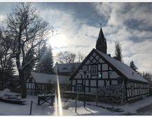 Fachwerkhäuser in einem verschneiten Dorf, beleuchtet von Sonnenlicht, das durch eine teils bewölkte Himmel bricht. Im Hintergrund ist ein Kirchturm mit spitzem Dach zu sehen, umgeben von kahlen Bäumen.