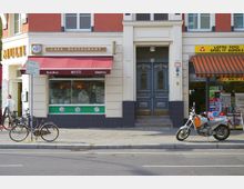 Straßenansicht mit einer Hausfassade, die ein Café-Restaurant mit roter Markise, eine blaue Eingangstür und einen Lottogeschäft mit orange-gestreifter Markise zeigt. Davor stehen ein Motorrad und zwei Fahrräder auf dem Gehweg.
