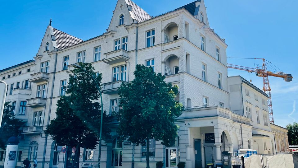 Großes, weißes mehrstöckiges Gebäude im Altbaustil mit Satteldächern, Giebeln, Balkonen und hohen Fenstern an einer Straßenecke. Vor dem Haus stehen Bäume; rechts sind ein Bauzaun und ein hoher Baukran vor einem angrenzenden Gebäudeteil zu sehen.