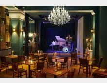 Gemütliche Bar mit einem kleinen Bühnenbereich, auf dem ein weißer Flügel und ein Schlagzeug stehen, umgeben von dunklen Vorhängen. Der Raum ist mit Holzmöbeln, Kerzenständern und einem großen Kristallleuchter dekoriert.