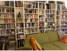 Ein Wohnzimmer mit einer großen, weißen Bücherwand, die bis zur Decke reicht und gefüllt ist mit Büchern, Dekoobjekten und Bildern. Im Vordergrund steht ein grünes Sofa mit orangefarbenen und grünen Kissen, daneben eine Stehlampe.