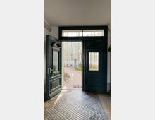 Geöffnete dunkle Holztüren in einem Gebäudeeingang, mit einem Blick auf einen gepflasterten Innenhof, umgeben von sandfarbenen Wohngebäuden. Der Boden besteht aus dunklen Fliesen, und im Hintergrund ist ein Fahrrad zu sehen.