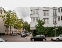 Wohnstraße mit mehrstöckigen Wohngebäuden, darunter ein Eckhaus mit hellem Anstrich, teils mit Efeu bewachsen. Pflastersteinstraße mit parkenden Autos entlang der Gehwege, flankiert von Bäumen und Straßenlaternen, bei bewölktem Himmel.