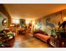 Raum im Vintage-Stil mit antiken Möbeln, darunter ein rotes Sofa, ein Frisiertisch mit Spiegeln und einem alten Stuhl. Die Wände sind mit gemalten Pfauen und goldener Verzierung dekoriert, der Holzboden und die warme Beleuchtung verstärken die nostalgische Atmosphäre.