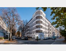 Eckgebäude im modernen Stil mit weißer Fassade und geschwungenen Balkonen, gelegen an einer Straßenkreuzung mit parkenden Autos und kahlen Bäumen im Herbst. Ein Fahrradfahrer fährt die Straße entlang, während vereinzelte Gebäude und Bäume die Umgebung prägen.