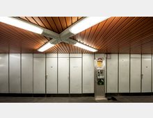 Ein U-Bahn-Bereich mit einer Wand aus hellgrauen Metallpaneelen, die mit nummerierten Türen versehen ist, sowie einer öffentlichen Telefonzelle auf der rechten Seite. Die Decke besteht aus braunen Holzpaneelen mit langen, hell erleuchteten Leuchtstofflampen.