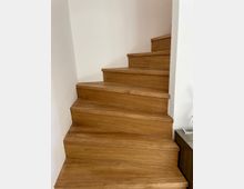 Holztreppe mit massivem braunem Holz und klaren Kanten, die an einer weißen Wand entlang nach oben führt. Im rechten Bildbereich ist ein Teil einer dunklen Holzkonsole sichtbar.