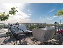 Dachterrasse mit Holzboden, modernen Gartenmöbeln, darunter Stühle aus Metall und ein Sofa mit weißen Kissen, sowie Pflanzen in Blumenkästen; im Hintergrund die Skyline einer Stadt mit dem Fernsehturm und klarem Himmel.