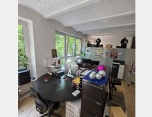 Helles Büro mit einem runden schwarzen Schreibtisch im Vordergrund, einem Computer und Büromaterialien. Im Hintergrund sind große Fenster mit Blick auf grüne Bäume, mehrere Schränke und dekorative Elemente wie glitzernde Kugeln sichtbar.