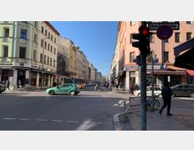 Straßenszene an einer Kreuzung mit mehrstöckigen Wohn- und Geschäftsgebäuden, darunter Restaurants und Geschäfte. Personen überqueren die Straße, Autos und ein Bus sind teilweise sichtbar, und im Vordergrund steht eine Ampel.