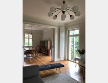 Wohnzimmer eines Hauses mit Parkettboden, einem grauen Sofa und einer gepolsterten Bank im Vordergrund. Durch eine große Türöffnung ist ein Esszimmer mit Holztisch und Stühlen sowie einem Holzschrank sichtbar, und rechts führen hohe Fenstertüren ins Grüne.