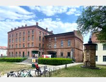 Backsteingebäude mit historischer Industriearchitektur, rechteckiger Grundform und Sprossenfenstern, flankiert von einem kleinen parkartigen Platz mit einer dekorativen, achteckigen Brunnenanlage im Vordergrund. Im Umfeld befinden sich Grünflächen, Fahrräder und moderne Reklametafeln.