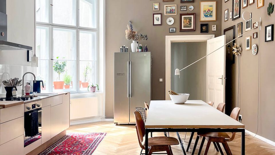 Bright open-plan kitchen and dining area with high ceilings, ornate ceiling molding, and large multi-pane windows. A long dining table sits on herringbone wood floors beside modern kitchen cabinets, a red runner rug, a stainless-steel fridge, and a gallery wall of framed artwork.