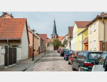 Gepflasterte Straße in einer Kleinstadt mit bunten Wohnhäusern auf beiden Seiten. Im Hintergrund ist ein Kirchturm mit Spitzdach zu sehen, umgeben von einigen Bäumen.