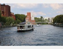 Ein Fluss mit zwei Booten, flankiert von einer Uferpromenade mit Bäumen und roten Backsteingebäuden links. Im Hintergrund eine Brücke, moderne Gebäude und ein Industriekomplex mit Türmen.