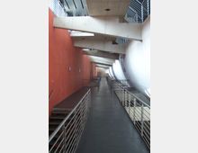 Ein moderner Innenbereich mit einem langen Gang, der von einer roten Wand auf der linken Seite und einer weißen geschwungenen Wand auf der rechten Seite flankiert wird. Sichtbare Metallgeländer und große Betonträger prägen die Architektur, während ein Boden aus dunklem Material den Gang abschließt.