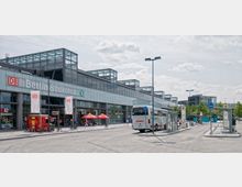 Der Eingang des Bahnhofs Berlin Südkreuz mit einer großen Glasfassade und dem Schriftzug des Bahnhofs und DB-Logos. Im Vordergrund befinden sich Bushaltestellen, ein parkender Reisebus, Fahrradständer und einige Verkaufsstände mit roten Sonnenschirmen.