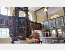 Innenraum einer historischen Kirche mit barockem Altar aus dunklem Holz, verziert mit goldenen Ornamenten, Figuren und christlichen Motiven. Im Hintergrund befinden sich Emporen mit hellblau-grauen Holzvertäfelungen und hohen Fenstern, die Tageslicht hereinlassen.