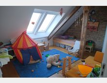 Kinderzimmer unter dem Dach mit schrägen Decken und zwei Dachfenstern, einem sichtbaren Holzbalken und einer Backsteinwand. Der Raum enthält ein Zelt in Rot-Gelb-Blau, ein niedriges Bett, Holzmöbel, Spielzeug und eine blaue Teppichfläche.