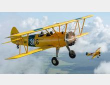 Zwei historische Doppeldecker-Flugzeuge im Flug über einer Landschaft mit Feldern, umgeben von weißen Wolken. Das vordere Flugzeug ist gelb mit der Beschriftung „US NAVY 326“ und einem sichtbaren radialen Sternmotor.