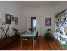 Wohnzimmer mit Holzboden und hohen Decken mit Stuckverzierungen. In der Mitte steht ein Esstisch mit Stühlen, links ein Sideboard mit Blumen und Kunstwerken darüber, rechts ein Bücherregal mit Pflanzen und Büchern, im Hintergrund eine Tür, die in einen anderen Raum führt.