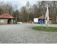Ein gepflasterter Kreisverkehr mit einer modernen Skulptur aus Metall in der Mitte, umgeben von zwei Gebäuden mit roten Ziegeldächern und Wänden aus Naturstein. Im Hintergrund ein Wald und ein Informationsschild mit verschiedenen Logos und Hinweisen.