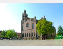 Historische Backsteinkirche mit Spitzenturm und rundem Rosettenfenster, umgeben von Bäumen und einem gepflasterten Platz; im Hintergrund sind Wohngebäude zu erkennen.
