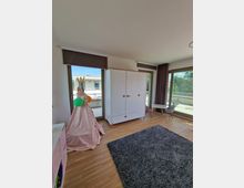 Innenraum eines hellen, modernen Kinderzimmers mit Holzfußboden. Es sind ein weißer Kleiderschrank, ein grauer Teppich, ein rosafarbenes Spielzelt mit Luftballons und große Fenster mit Blick auf einen Balkon zu sehen.