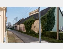 Schmale Dorfstraße mit einfachen Gebäuden, darunter eine alte Lagerhalle mit einem begrünten Dach und von Efeu bewachsender Wand. Ein Straßenschild mit der Aufschrift „Am Musterplatz“ ist sichtbar, umgeben von ländlicher Vegetation und vereinzelten Strommasten.