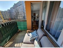 Ein kleiner Balkon mit grauem Sofa und Kissen, umgeben von einem grünen Sichtschutz. Im Hintergrund sind Wohngebäude und Bäume zu sehen, während eine geöffnete Glastür ins angrenzende Wohnzimmer mit Esstisch und Hängelampe führt.