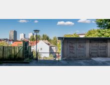 Städtische Szene mit grauen Garagen, einigen von Graffiti bedeckt, im Vordergrund. Im Hintergrund sind Wohnhäuser mit roten Dächern, Bäume und hohe moderne Gebäude unter einem blauen Himmel mit weißen Wolken zu sehen.