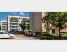 Wohnsiedlung mit mehrstöckigen, farbig gestrichenen Wohngebäuden, einigen Balkonen und Grünflächen im Vordergrund. Parkende Autos und ein gepflasterter Weg sind zwischen den Gebäuden zu sehen, umgeben von Bäumen.