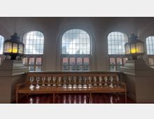 Großer Innenraum mit hohen Rundbogenfenstern, die Tageslicht durchlassen. Im Vordergrund steht eine Holzbank zwischen zwei verzierten Laternen auf steinernen Sockeln.