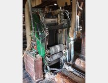 Alte Holzbearbeitungsmaschine in einer Werkstatt mit sichtbaren Zahnrädern, Schläuchen und Holzresten auf dem Boden. Im Hintergrund sind weitere Werkzeuge und Holzstapel zu sehen.