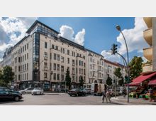 Straßenszene in einer Stadt mit einem mehrstöckigen Wohn- und Geschäftsgebäude im Vordergrund, das helle Fassaden mit großen Fenstern hat. Im unteren Bereich des Gebäudes befinden sich Geschäfte, davor geparkte Autos und ein Obststand, mit Ampeln und Fußgängern im Straßengeschehen.