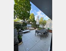 Terrasse mit großen, hellen Steinfliesen und moderner Einrichtung, darunter ein langer schwarzer Tisch mit Stühlen und eine Liege. Im Hintergrund ein Gartenbereich mit Bäumen, Sträuchern, einem Sonnenschirm und Kiesflächen, umgeben von Nachbarhäusern.