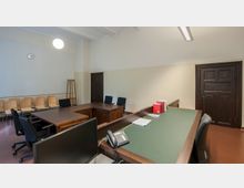 Ein schlicht eingerichteter Büroraum mit hellen Wänden, dunklen Holztüren und mehreren Holztischen. Im Raum befinden sich Stühle, ein Telefon, Aktenordner sowie ein Computerbildschirm, und an der Wand hängt eine Uhr.