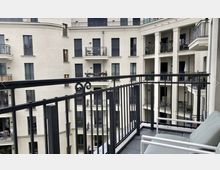 Blick von einem Balkon auf einen modernen Innenhof eines Wohngebäudes mit mehreren Etagen. Die Fassade ist in hellen Farben gehalten, mit Balkonen, verzierten Eisengeländern und regelmäßig angeordneten Fenstern.