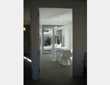 Ein moderner, minimalistisch eingerichteter Essbereich mit einem weißen Tisch und geschwungenen weißen Stühlen. Im Hintergrund befindet sich eine Glastür, die auf eine Terrasse mit blühendem Baum führt und viel Tageslicht in den Raum lässt.