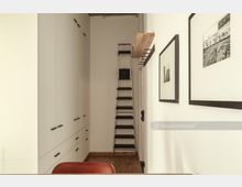 Schmaler Innenraum mit weißem Einbauschrank auf der linken Seite, einer Trittleiter in der Mitte, einem Holzregal an der Wand und gerahmten Schwarz-Weiß-Fotografien auf der rechten Seite. Der Boden besteht aus Holz, und der Raum hat eine minimalistische Gestaltung.