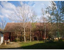 Eine große Backsteinscheune mit einem roten Ziegeldach, umgeben von kahlen und vereinzelt belaubten Bäumen. Im Vordergrund ist ein kleiner Hof mit Grasfläche, losen Gegenständen und einem Teil eines weißen Nebengebäudes sichtbar.