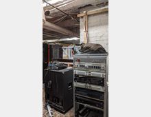 Kellerraum mit technischer Ausstattung, einschließlich eines Serverschranks mit Audiogeräten und einem Lautsprecherkasten mit Kabeln. Die weißen Ziegelwände und freiliegenden Rohre sowie Kabel an der Decke betonen die einfache, industrielle Umgebung.