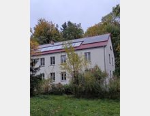 Zweistöckiges Wohnhaus mit weißer Fassade, rotem Ziegeldach und installierten Solarpaneelen, umgeben von dichtem Grün und Bäumen im Herbst. Im Vordergrund befindet sich eine grasbewachsene Fläche mit vereinzeltem Laub.