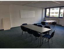 Ein einfacher Besprechungsraum mit grauem Teppichboden, zwei rechteckigen Tischen, umgeben von schwarzen Stühlen, und einem großen Fenster, das Tageslicht von außen hereinlässt. An der weißen Wand hängt ein leeres Whiteboard, und der Raum wirkt funktional und schlicht eingerichtet.