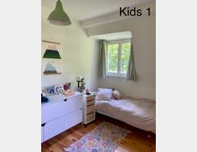 Ein helles Kinderzimmer mit Holzdielenboden, Einzelbett mit heller Bettwäsche, einem weißen Sideboard mit Deko und Fotos sowie ein kleines Rollcontainer daneben. Ein Fenster mit grünen Vorhängen lässt Tageslicht herein, und an der Wand hängt ein Poster mit Bergmotiven.