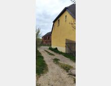 Ein schmaler, unbefestigter Weg führt zwischen einem gelben Wohnhaus mit Satteldach und einem Holzzaun vorbei. Im Hintergrund steht ein rotes Backsteingebäude mit mehreren Rundbogenfenstern, umgeben von Bäumen und Sträuchern.