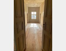 Blick in einen leeren Raum mit Holzdielenboden und unverputzten Wänden, der durch eine geöffnete Doppeltür sichtbar ist. Im Hintergrund befindet sich ein Fenster, durch das Tageslicht einfällt; die Decke zeigt Spuren von Abnutzung.